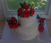 Here are edible, handmade Red Roses on top of two tiered white cake.  The cake has delicate scroll work from royal icing.  there are a dozen red roses.